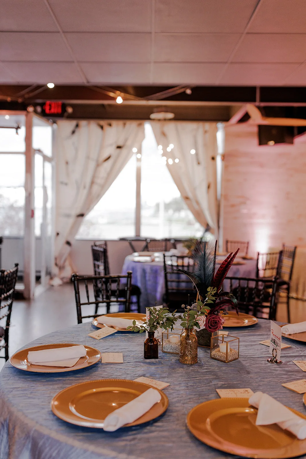 The Club set for a wedding reception with round tables, gold chargers and Chiavari chairs, the water in the windows beyond