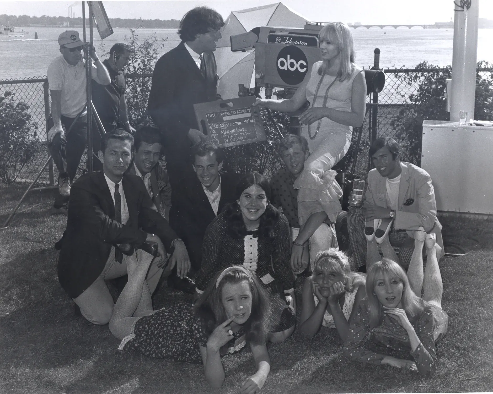A television crew and a group of young people gathered on the lawn at the river's edge, an ABC camera behind them and a woman holding up a chalked production slate.