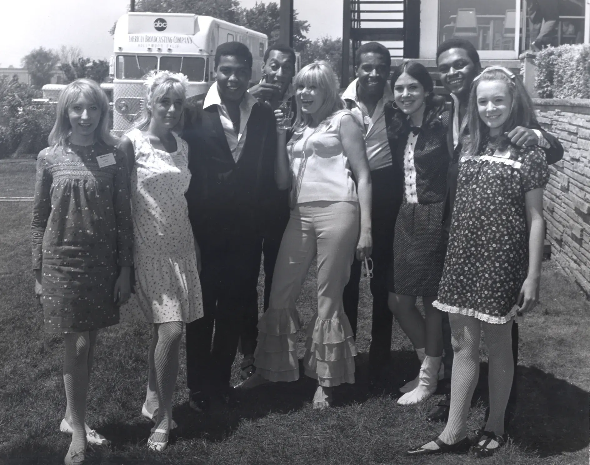 A group of young people posed shoulder to shoulder on the lawn, a large network mobile television unit parked on the drive behind them.