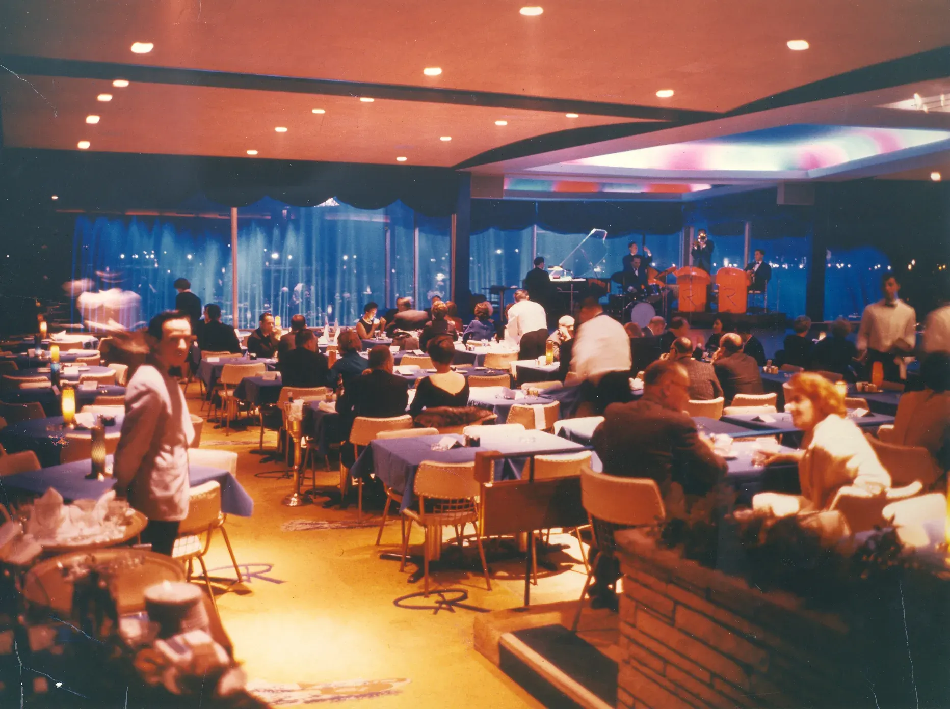 The Roostertail's main room at night, every table taken under warm ceiling light, a band playing at the far end against a wall of deep blue windows.