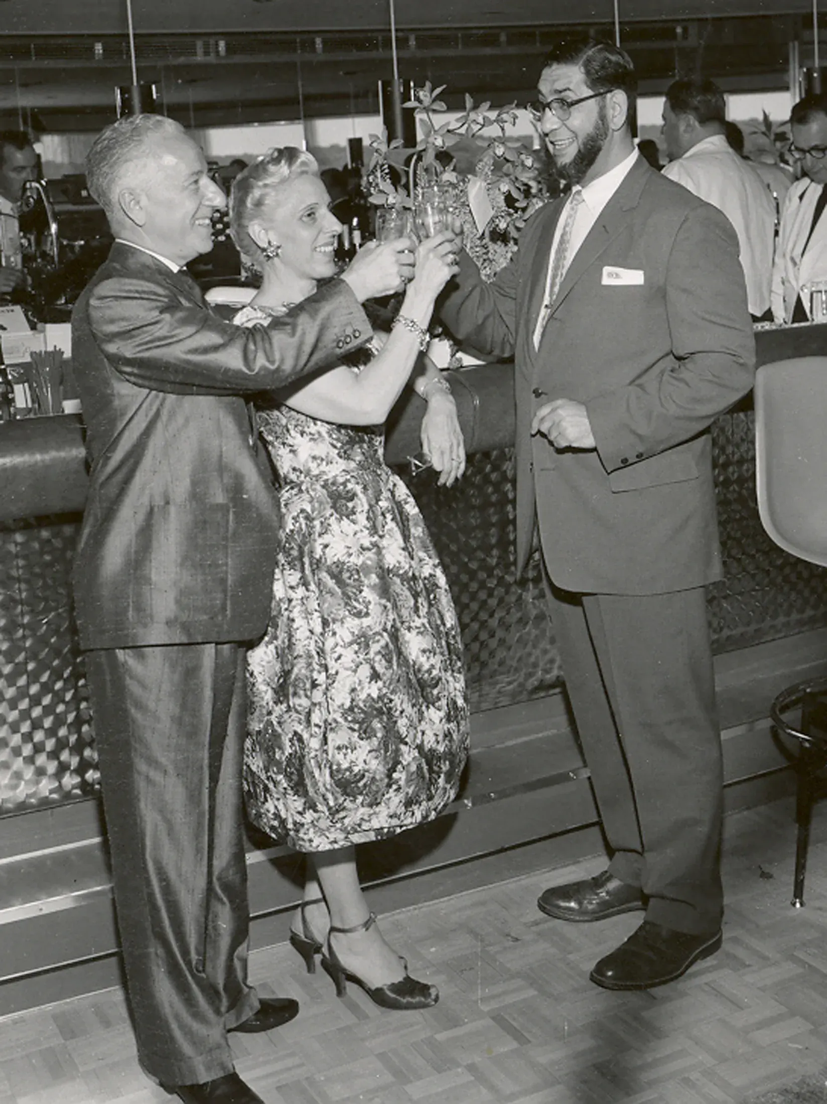 Three guests in midcentury evening dress lift tall glasses trimmed with orchid sprays, one woman wearing a corsage at her waist.