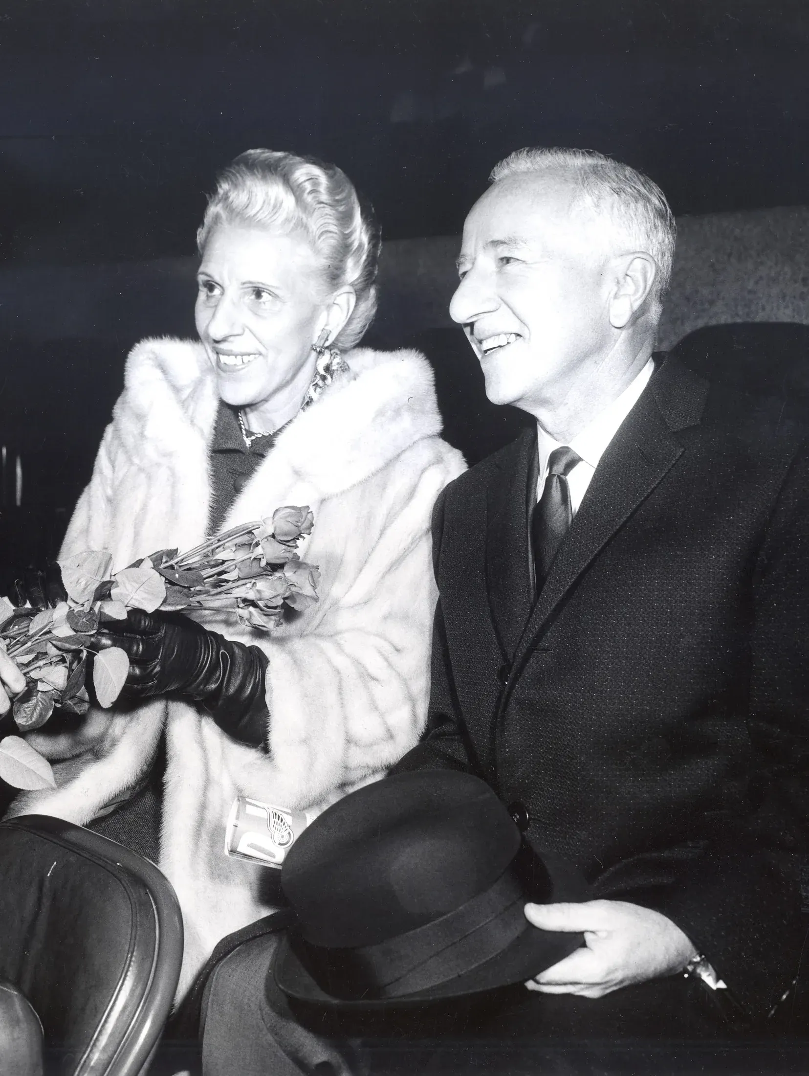 Joe and Millie Schoenith seated side by side in evening dress, she in a pale fur holding a bundle of roses, both smiling.