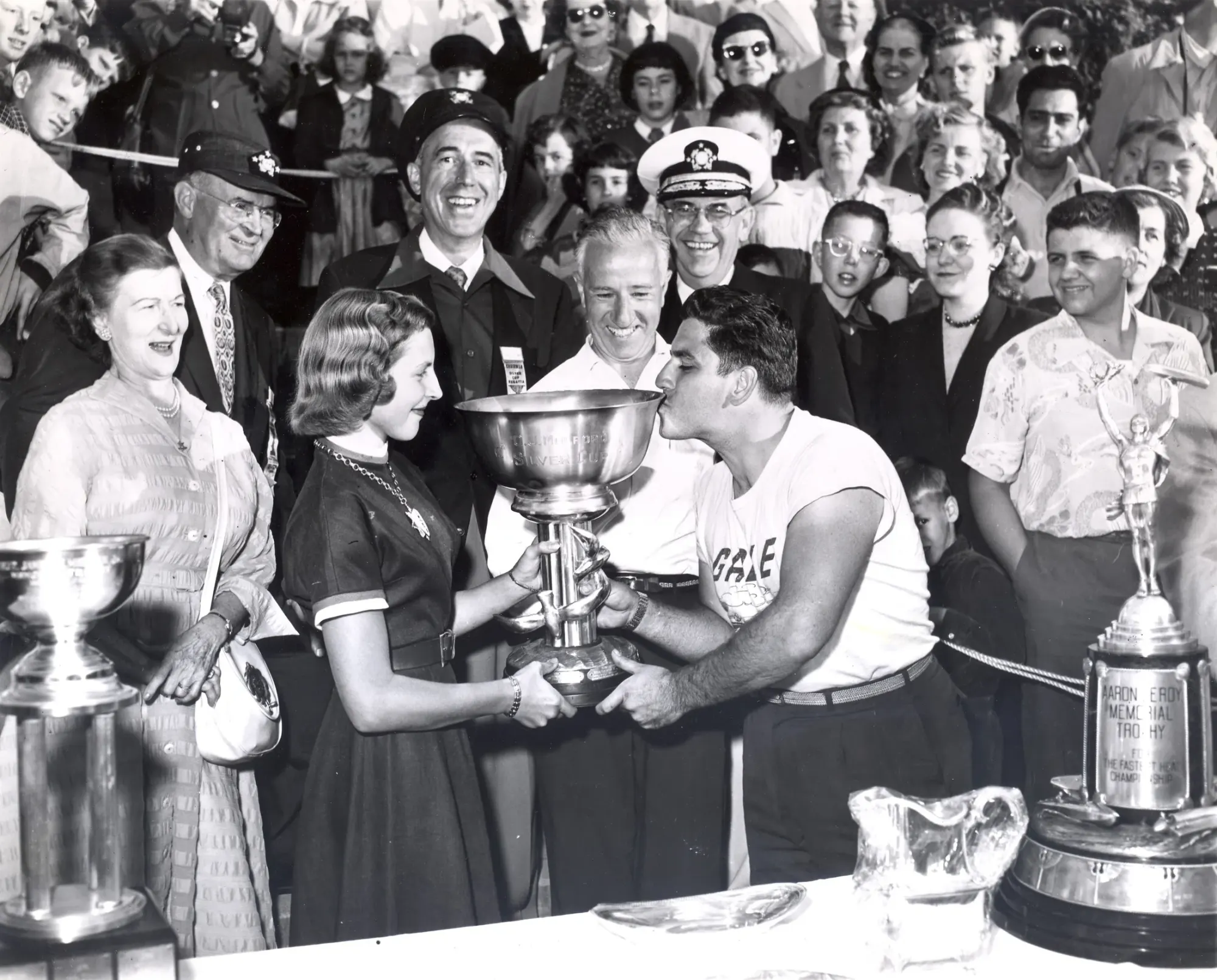 A young man in a GALE team shirt leans across a table and kisses the rim of a large silver trophy bowl held by a young woman, with a packed grandstand crowd, a police officer and a naval officer behind them.