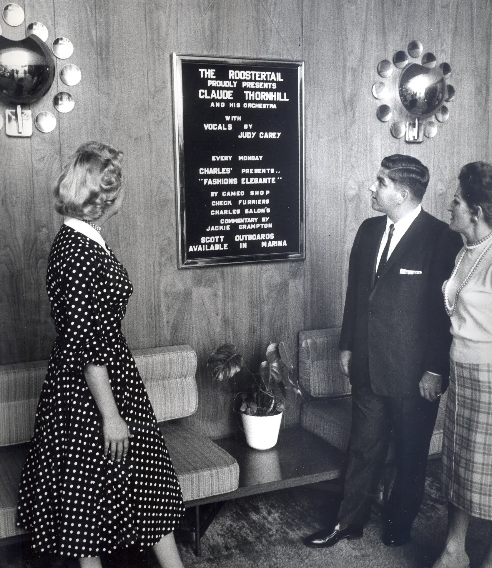 Three guests in evening clothes stand in a wood-paneled lobby reading a framed black letter board that lists the night's orchestra, a Monday fashion show, and outboards available in the marina.