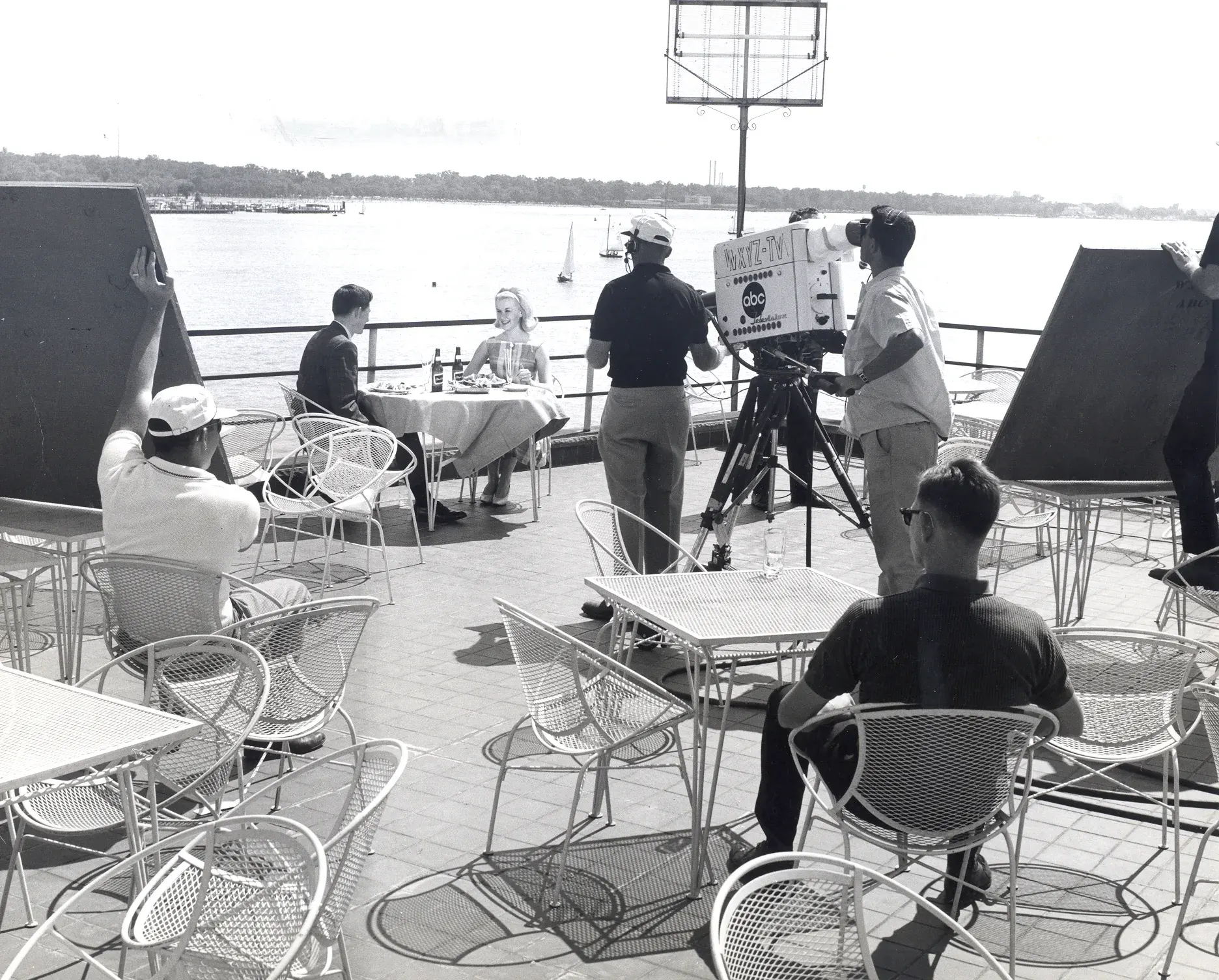 A wide view of the riverside terrace during a shoot, a television camera trained on a couple seated at a single table with the river and far shore behind them.