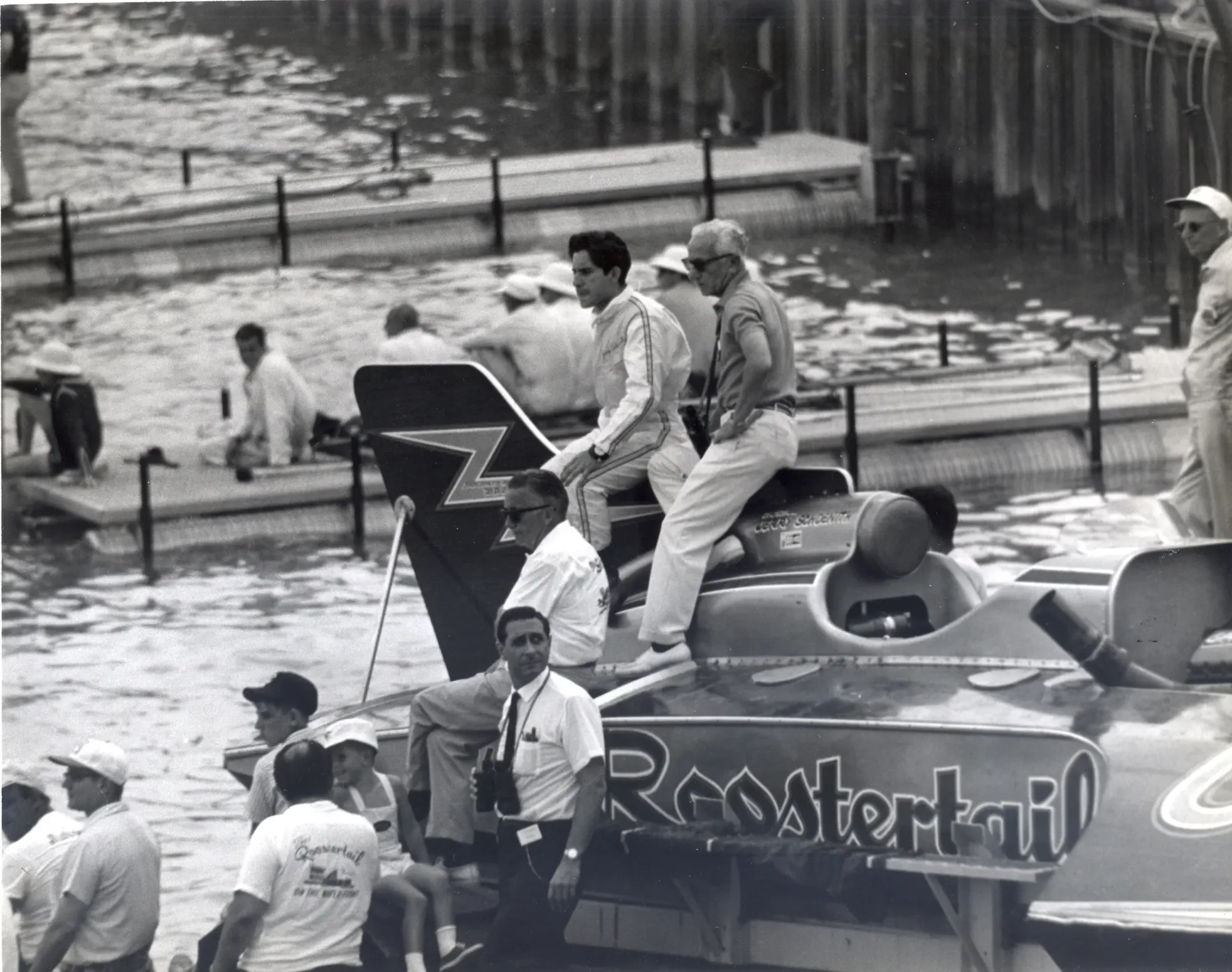 An unlimited hydroplane alongside a dock with crew standing on its deck, the word Roostertail lettered in script across the flank of the hull.