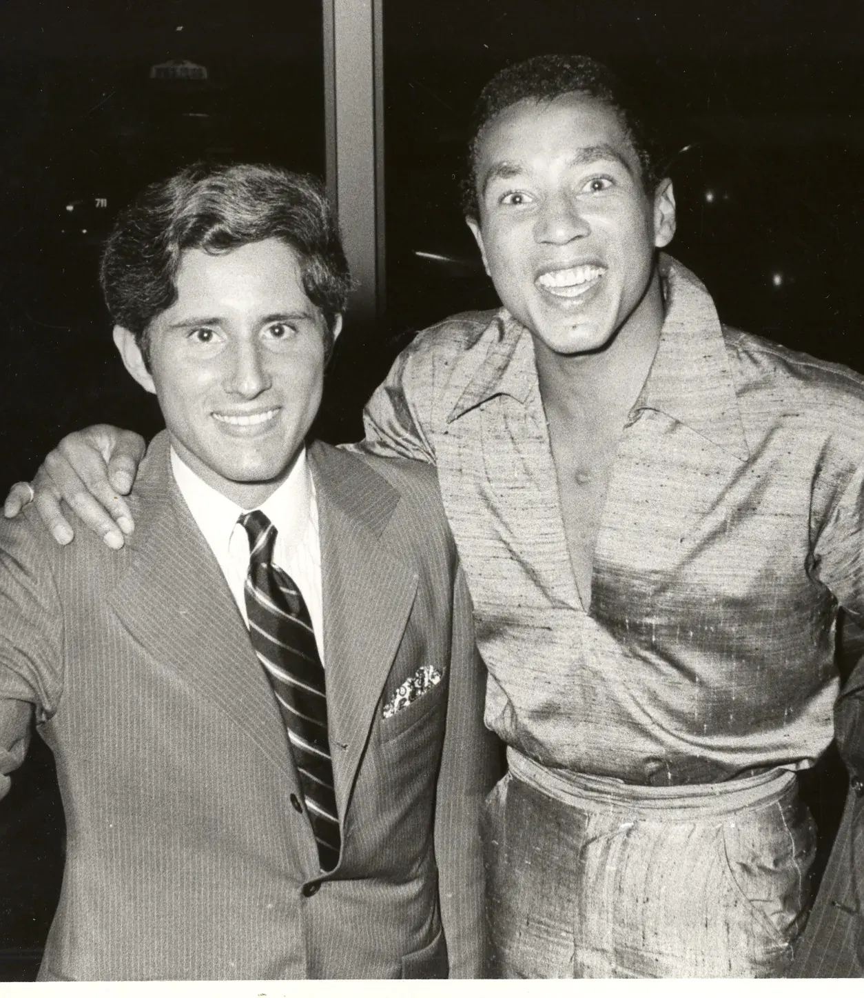Smokey Robinson with his arm around a younger man in a pinstriped suit and tie, both smiling at the camera, night windows behind them.